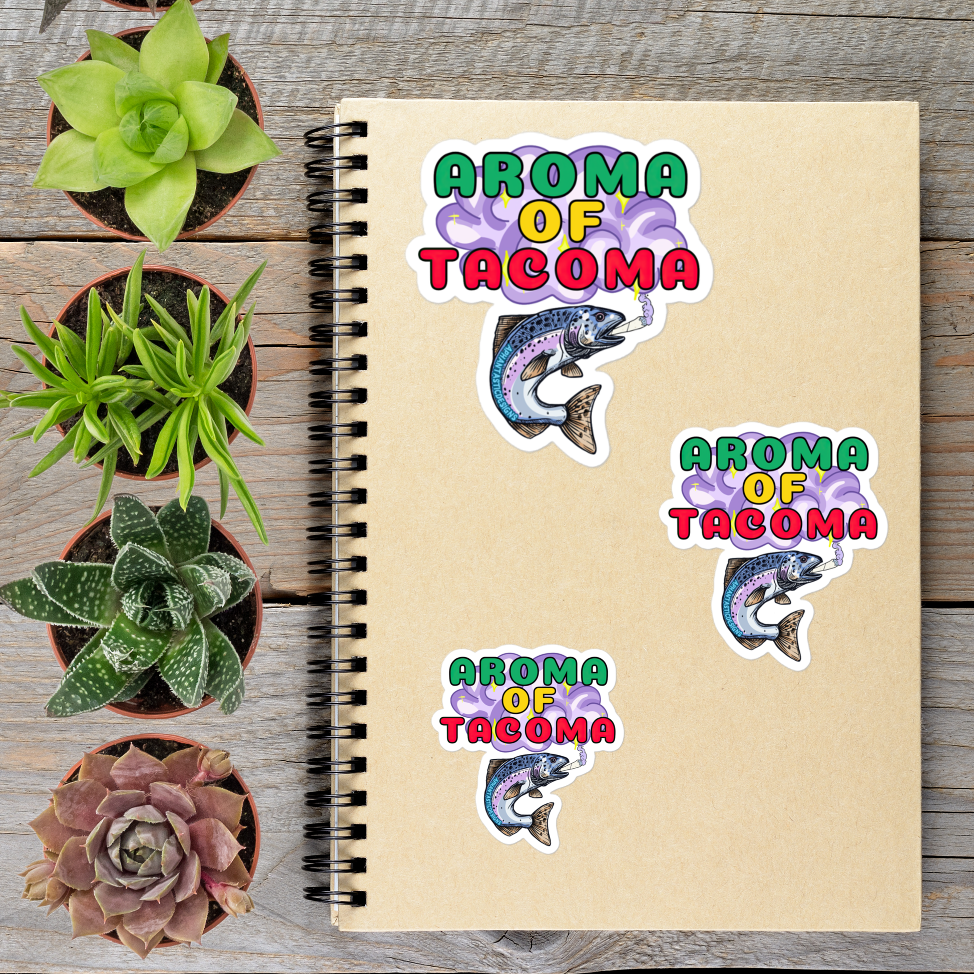 a spiral notebook with stickers on it, surrounded by potted plants on a wooden surface.