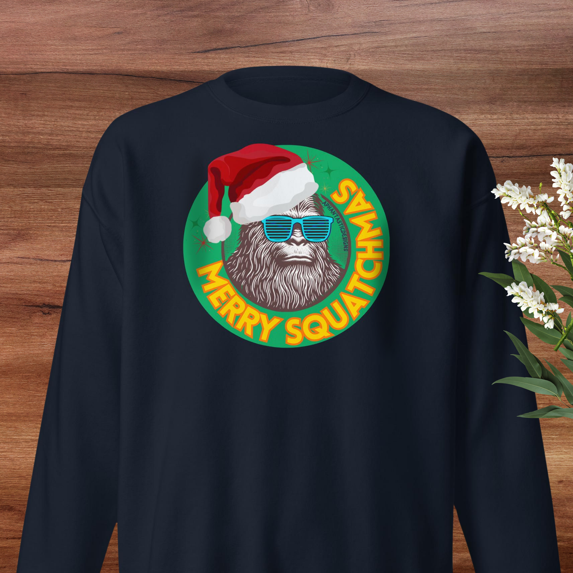 A black sweater with a graphic design featuring a smiling Sasquatch wearing a Santa hat and sunglasses, surrounded by the text "MERRY SQUATCHAT".