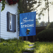 A blue flag with the text "Spokane Strong" hangs on a metal pole in a grassy yard, with a house and trees in the background.