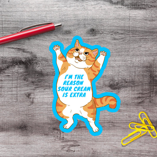 A sticker of a cat with a humorous caption, "I'm the reason sour cream is extra", is placed on a wooden surface next to a red pen and yellow paper clips.