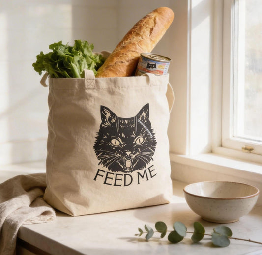 A beige tote bag with a cat face design and the text "FEED ME" sits on a kitchen counter, holding a loaf of bread and a can of tuna.