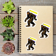 a spiral notebook with stickers of three Sasquatch-like creatures, each with a flag, placed on a wooden surface alongside several potted plants.