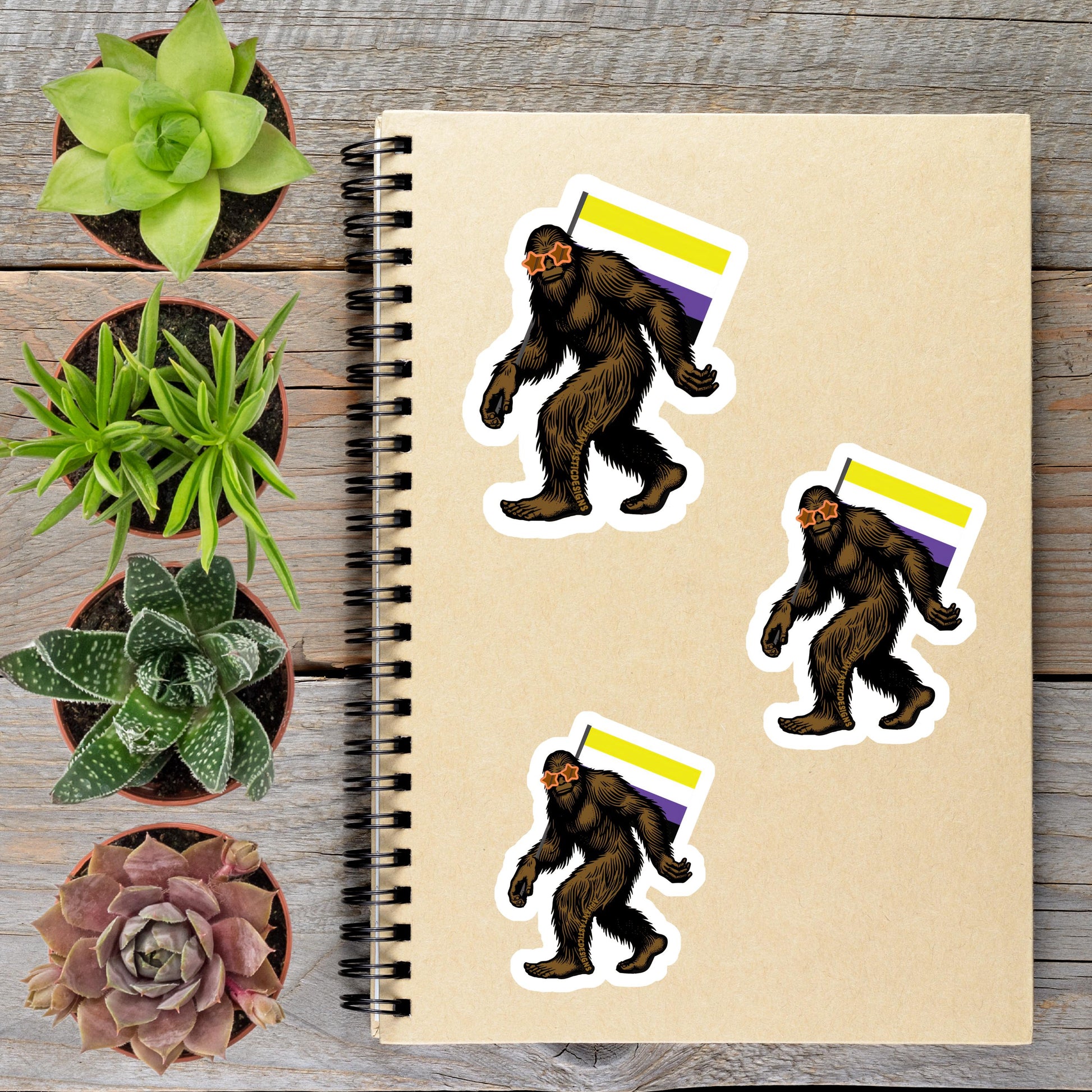 a spiral notebook with stickers of three Sasquatch-like creatures, each with a flag, placed on a wooden surface alongside several potted plants.