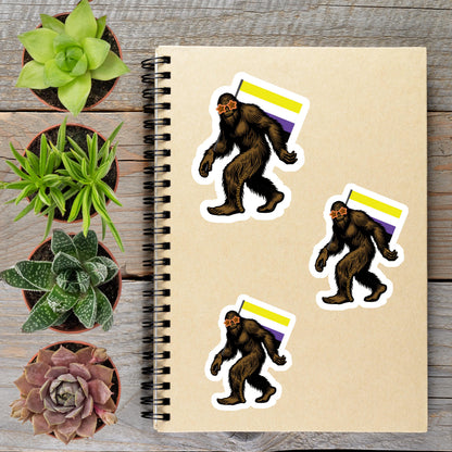 a spiral notebook with stickers of three Sasquatch-like creatures, each with a flag, placed on a wooden surface alongside several potted plants.