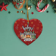A red heart-shaped ornament with the text "baby's first christmas 2026" printed on it, surrounded by Christmas decorations such as stars, candy canes, and pine branches.