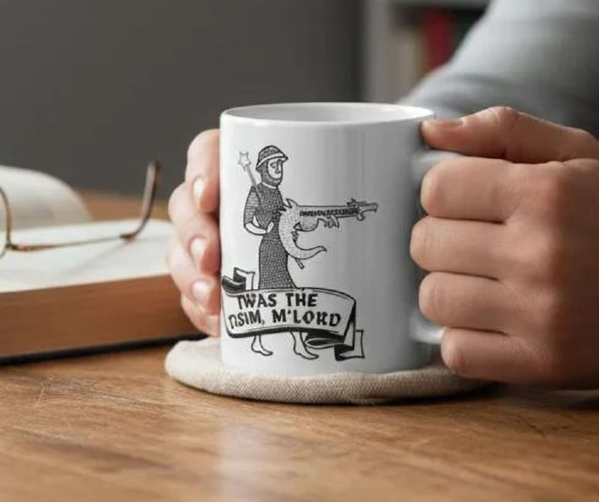 A person's hands holding a white mug with a black and white illustration of a person holding a gun and the text "TWAS THE ISIM, M LORD" printed on it.