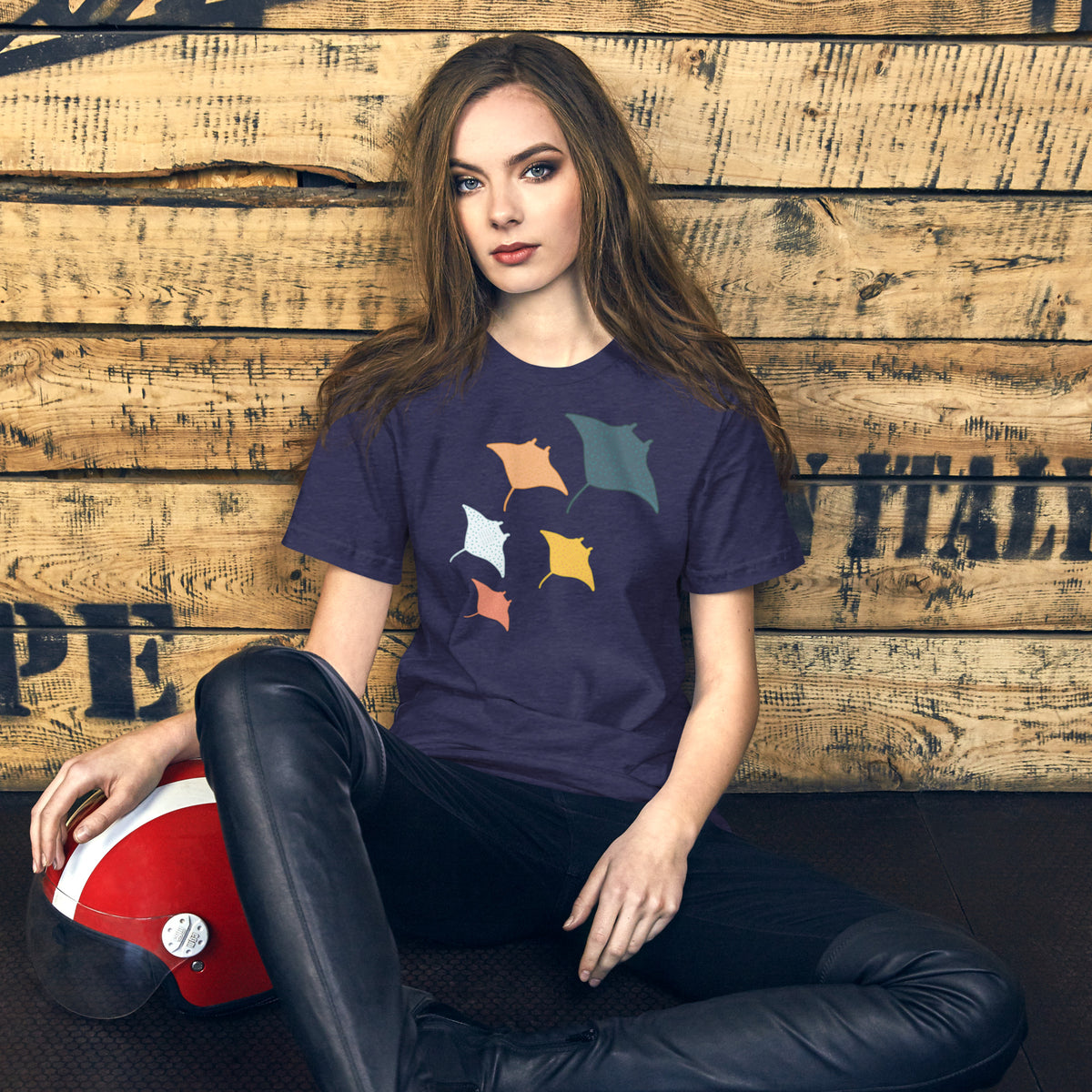 A young woman with long brown hair is sitting on the ground in front of a wooden wall, wearing a purple t-shirt with a graphic design.