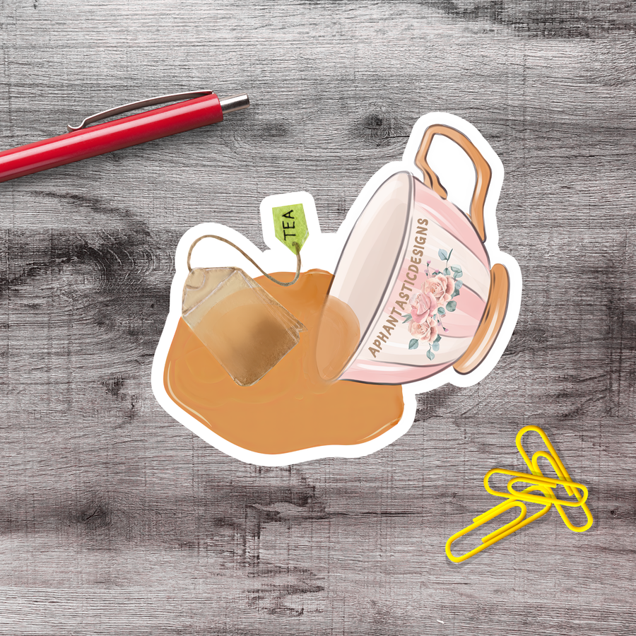A sticker depicting a teacup spilling tea, a pen, and a yellow stapler on a wooden surface.