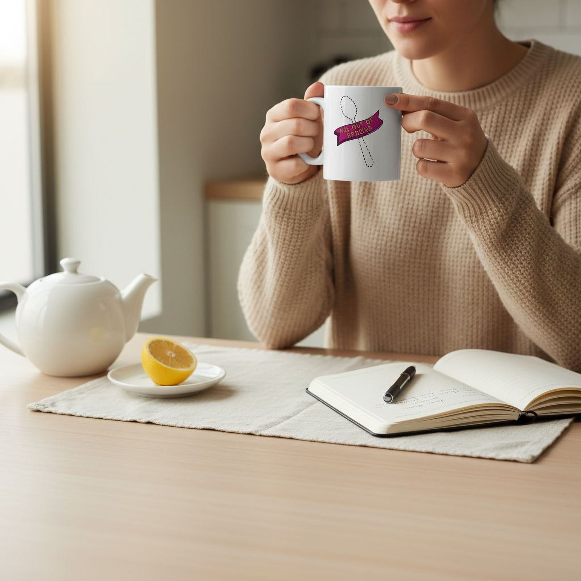A person is sitting at a table, holding a mug with a design on it, and there is a teapot, a plate with a lemon slice, and an open notebook on the table.