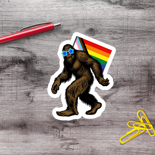 a sticker of a cartoon Sasquatch wearing sunglasses and carrying a rainbow flag, placed on a wooden surface next to a red pen and a pile of yellow paper clips.