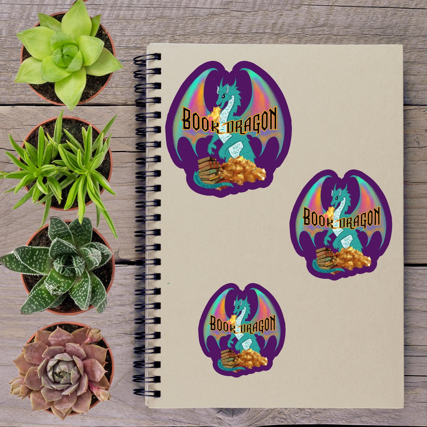 a spiral notebook with stickers of a dragon and a plant, along with several potted succulents arranged on a wooden surface.
