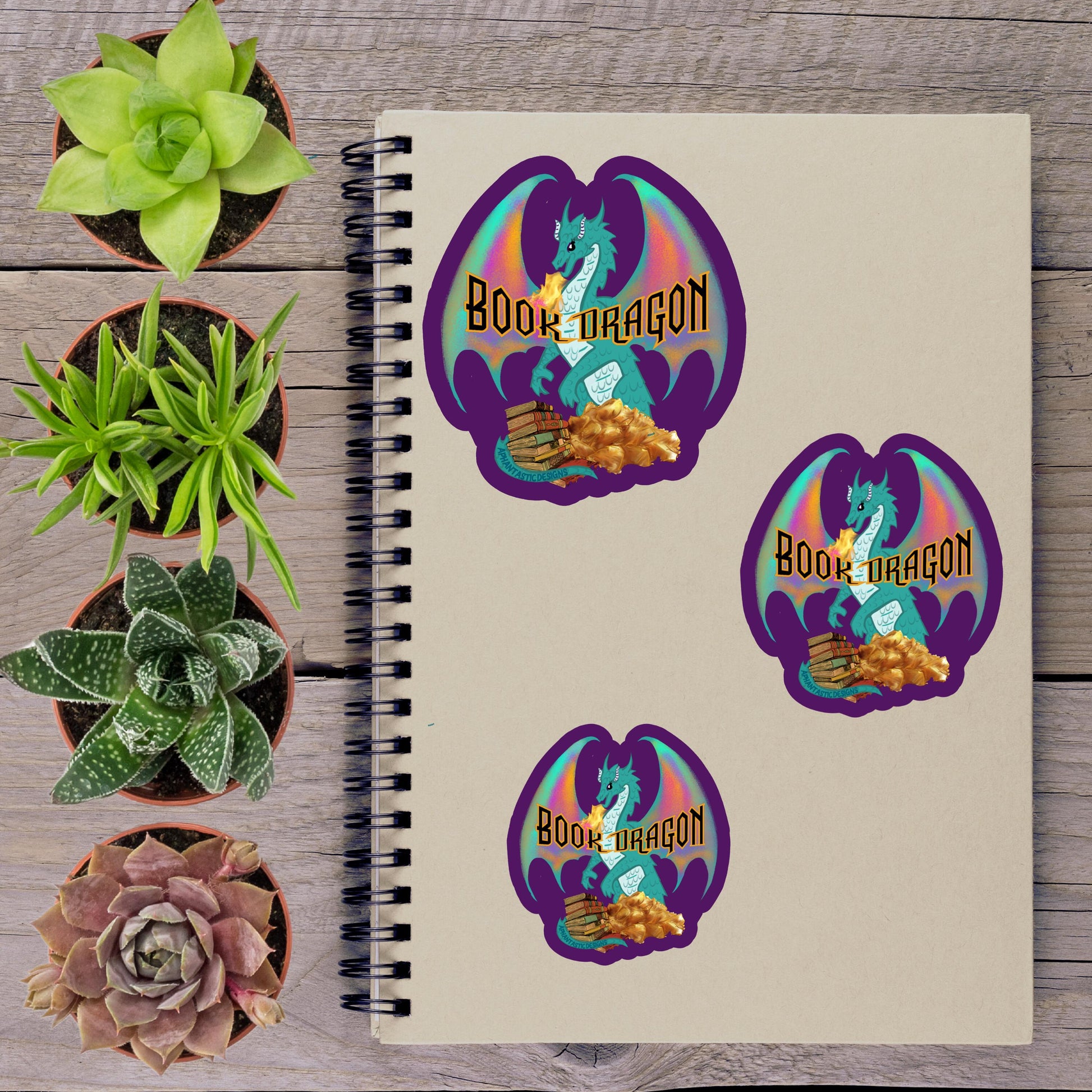 a spiral notebook with stickers of a dragon and a plant, along with several potted succulents arranged on a wooden surface.