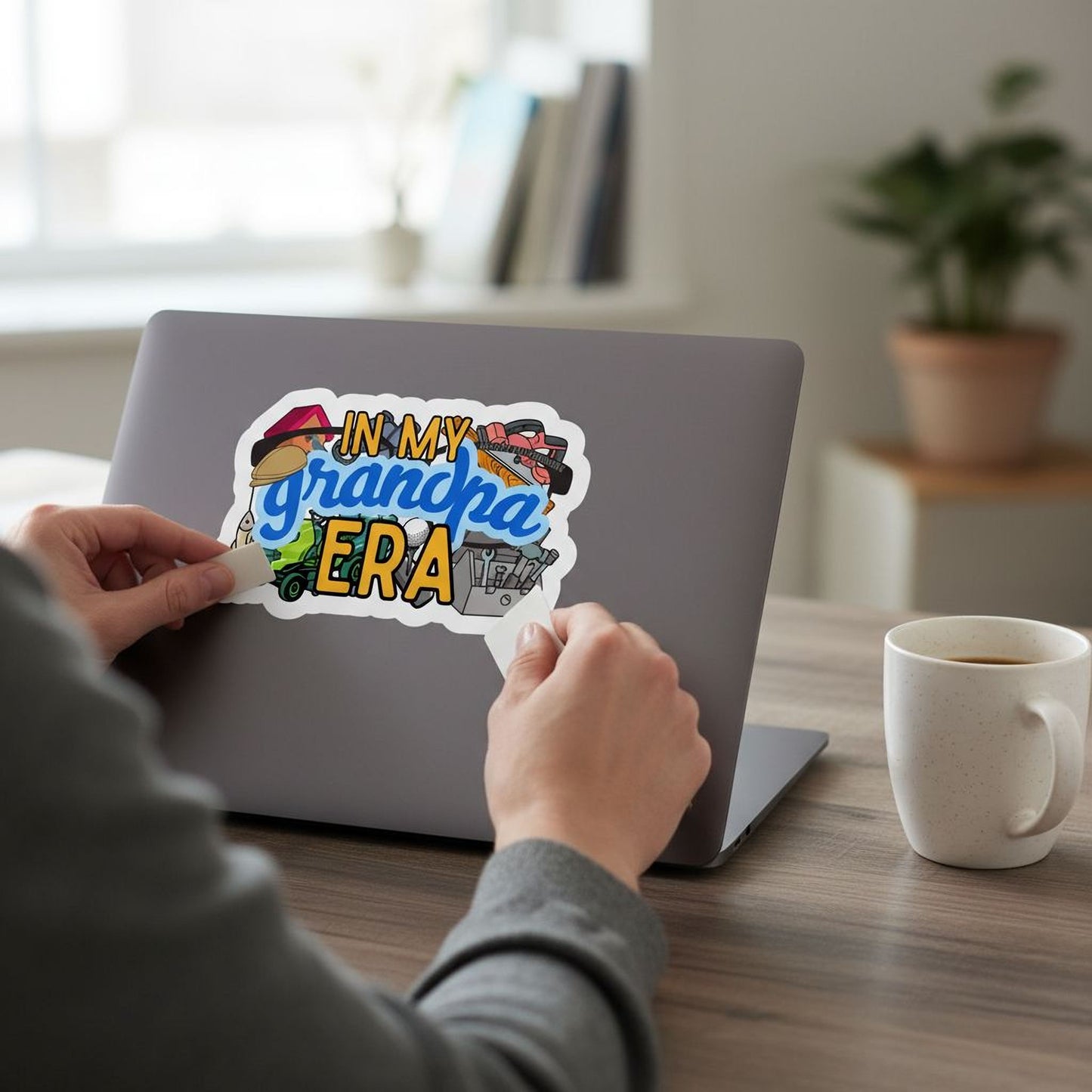 A person is sitting at a desk, working on a laptop with a sticker on the back. The sticker features a colorful design and text that says "In my grandpa era".