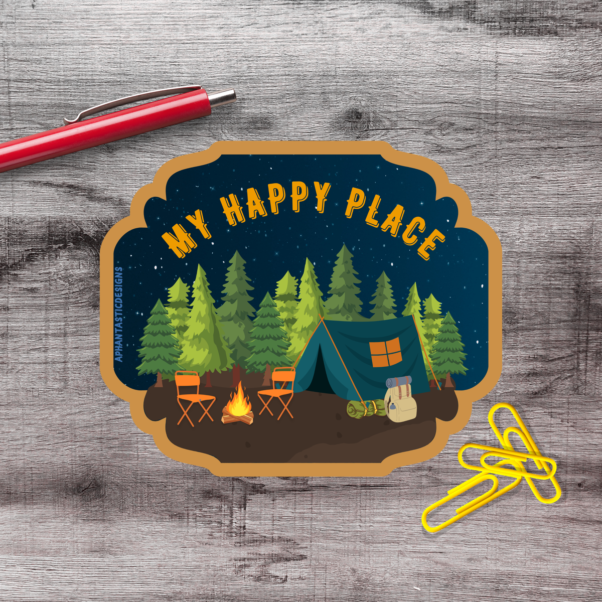 a sticker with a camping scene, including a tent, campfire, and chairs, along with a pen and a yellow stapler on a wooden surface.