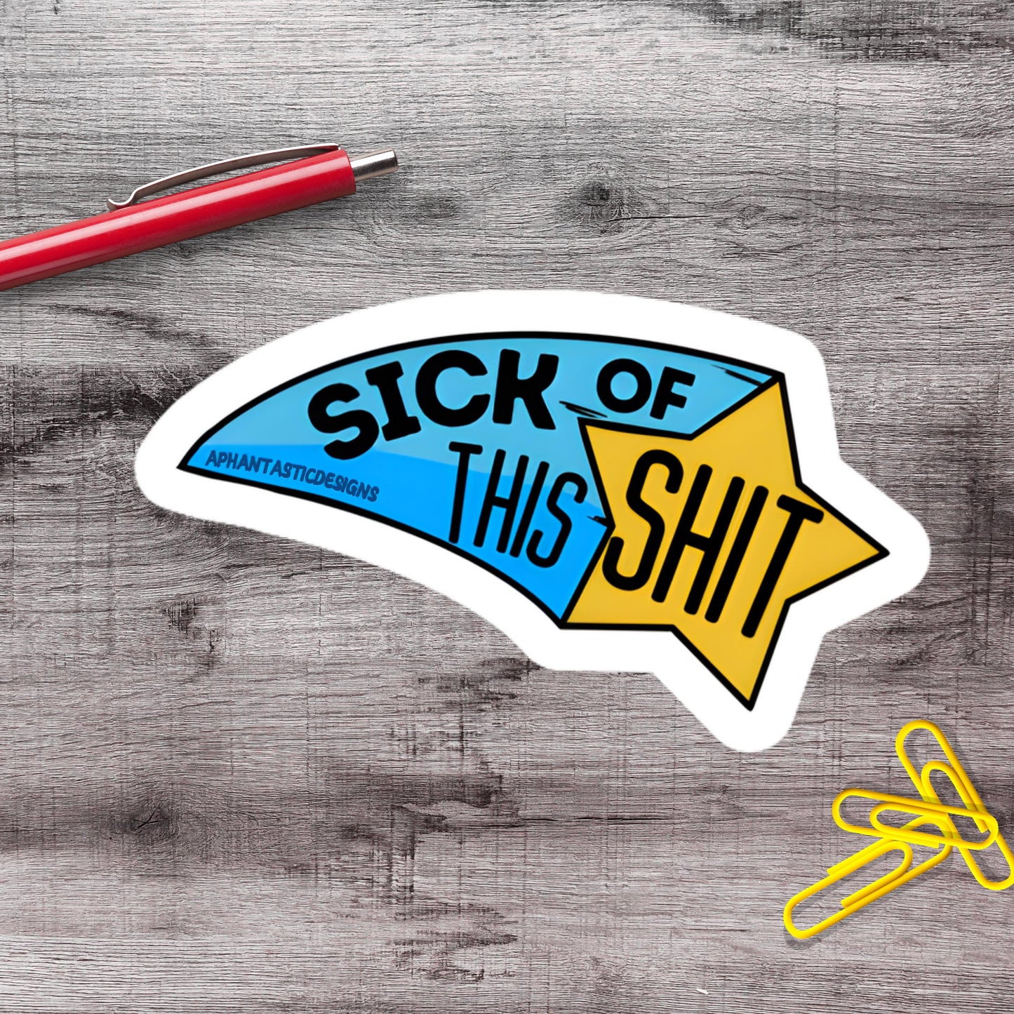 a sticker with the text "SICK OF THIS SHIT" and a star-shaped design, placed on a wooden surface alongside a red pen and yellow paper clips.
