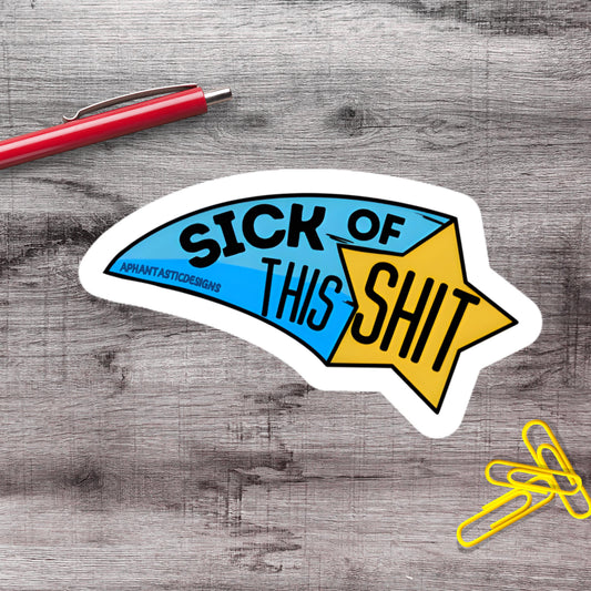 a sticker with the text "SICK OF THIS SHIT" and a star-shaped design, placed on a wooden surface alongside a red pen and yellow paper clips.