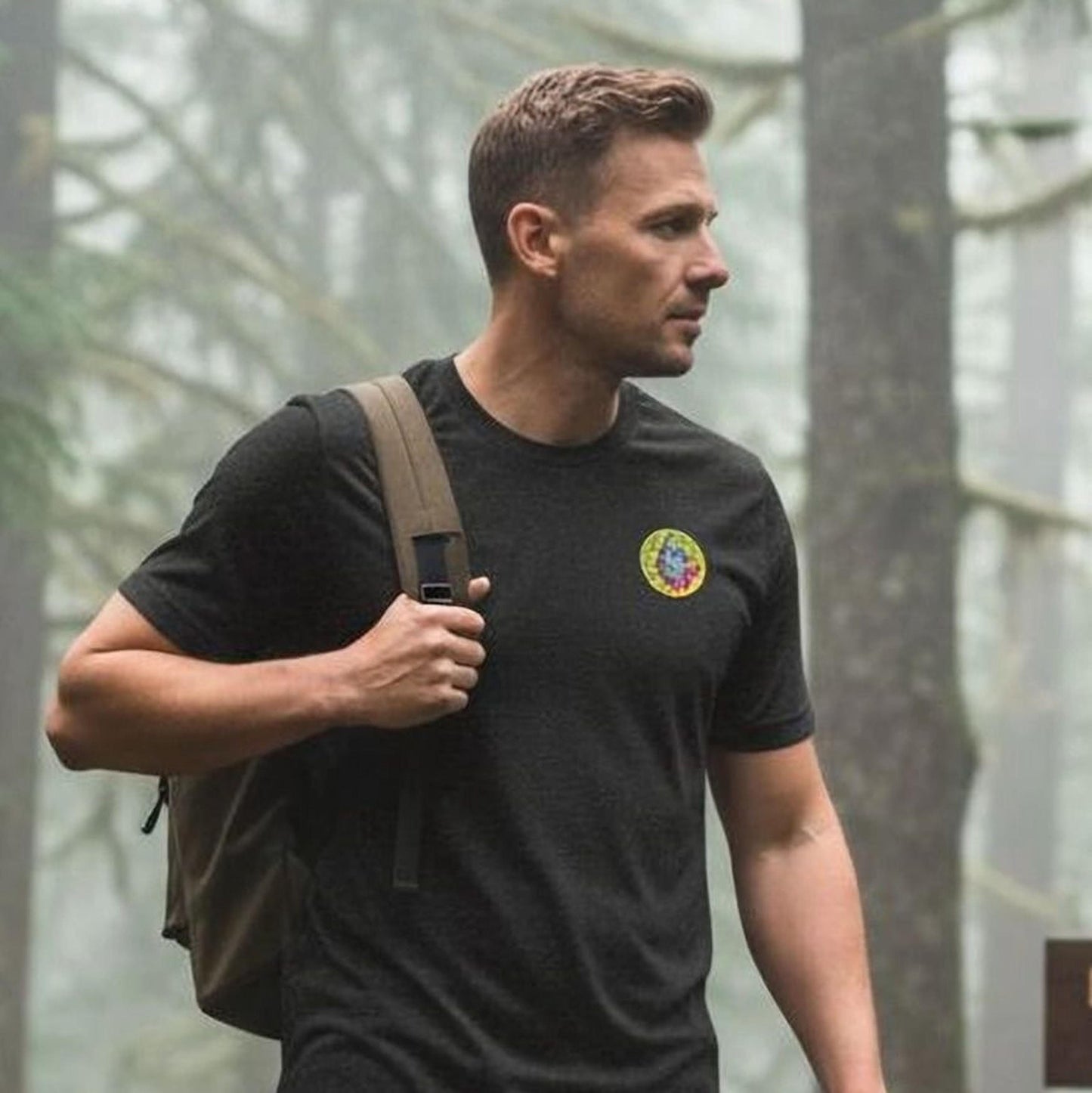 A man in a black shirt and jeans stands on a trail in a forest, holding a map and looking off to the side.
