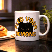 a mug with the text "FEED YOUR DEMONS" and a cinnamon roll on a kitchen counter, with a potted plant and a lamp in the background.