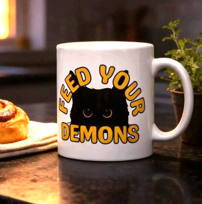 a mug with the text "FEED YOUR DEMONS" and a cinnamon roll on a kitchen counter, with a potted plant and a lamp in the background.