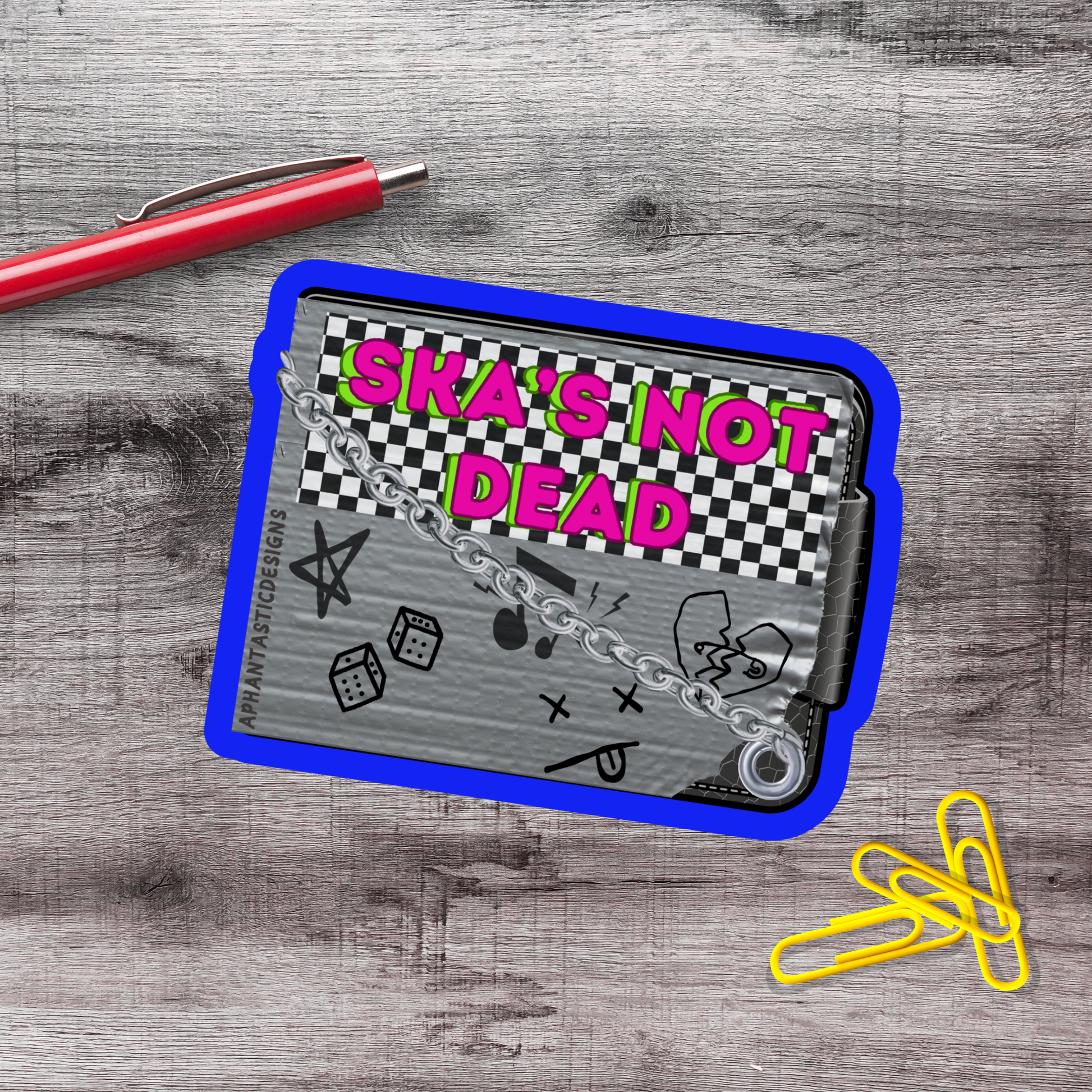 a sticker or decal with the text "Ska's not dead" on a blue and gray background, along with various graphic elements such as a checkerboard pattern, stars, and a broken heart. There is also a red pen and a few yellow paper clips on a wooden surface.