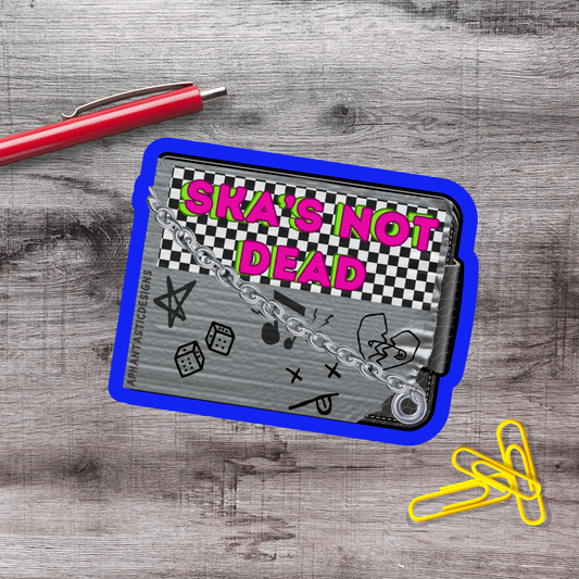 a sticker or decal with the text "Ska's not dead" on a blue and gray background, along with various graphic elements such as a checkerboard pattern, stars, and a broken heart. There is also a red pen and a few yellow paper clips on a wooden surface.