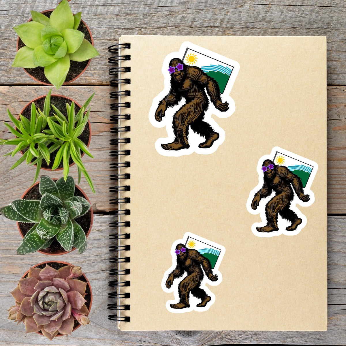 a spiral-bound notebook with stickers of cartoon-like Sasquatch figures, surrounded by potted plants and a wooden surface.