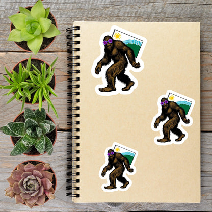 a spiral-bound notebook with stickers of cartoon-like Sasquatch figures, surrounded by potted plants and a wooden surface.