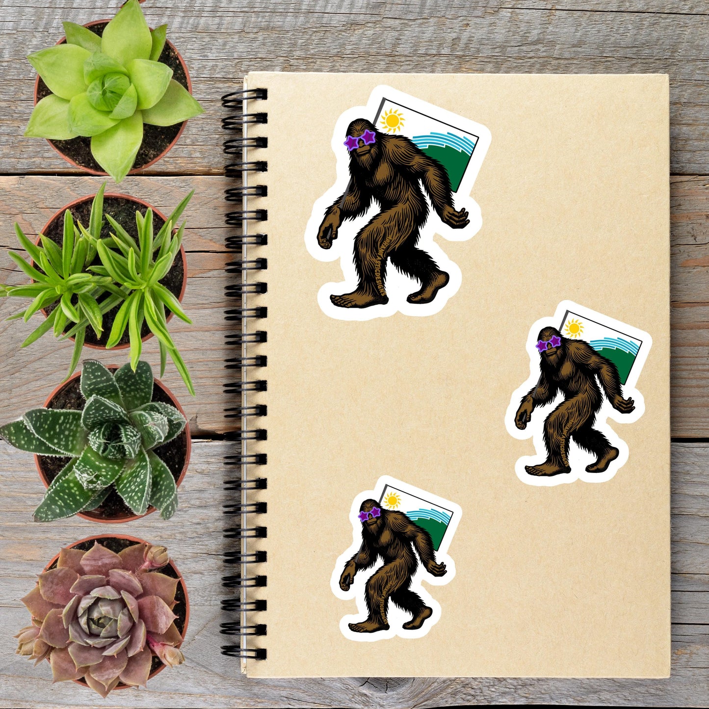 a spiral-bound notebook with stickers of cartoon-like Sasquatch figures, surrounded by potted plants and a wooden surface.