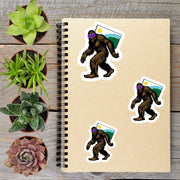 a spiral-bound notebook with stickers of cartoon-like Sasquatch figures, surrounded by potted plants and a wooden surface.