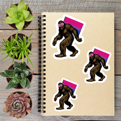 a spiral notebook with stickers of three cartoon-like Sasquatch figures wearing pink sunglasses, placed on a wooden surface alongside several potted plants.