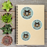 a spiral notebook with three circular stickers on it, each featuring a cat's face and the text "I'm into cats & tats". The stickers are placed on a wooden surface alongside several potted plants, creating a casual and relaxed atmosphere.