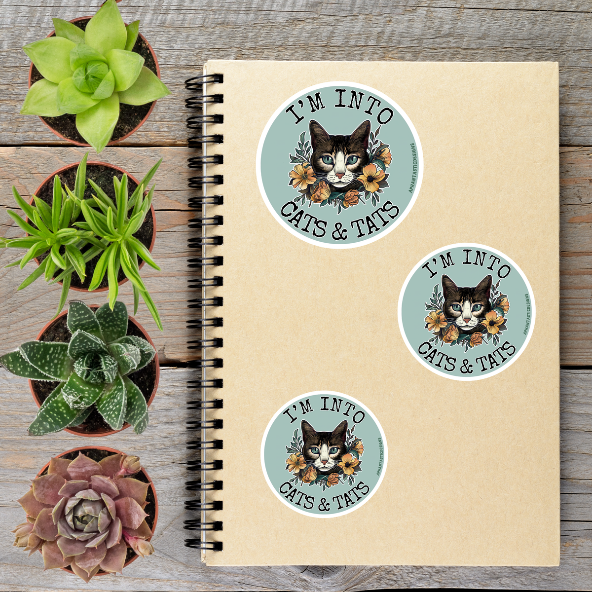 a spiral notebook with three circular stickers on it, each featuring a cat's face and the text "I'm into cats & tats". The stickers are placed on a wooden surface alongside several potted plants, creating a casual and relaxed atmosphere.