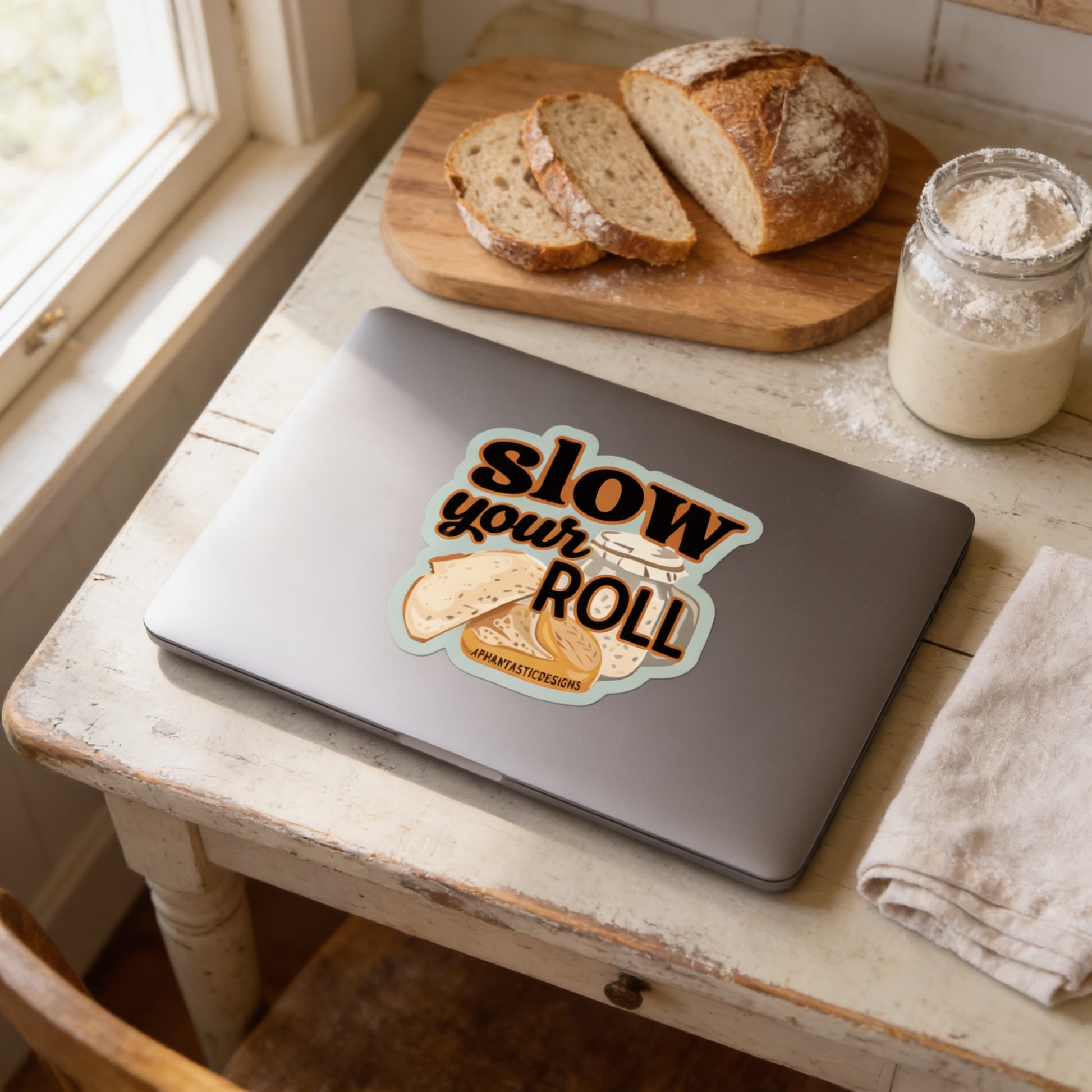 a laptop with a sticker on it, placed on a wooden table next to a loaf of bread and a jar of flour.