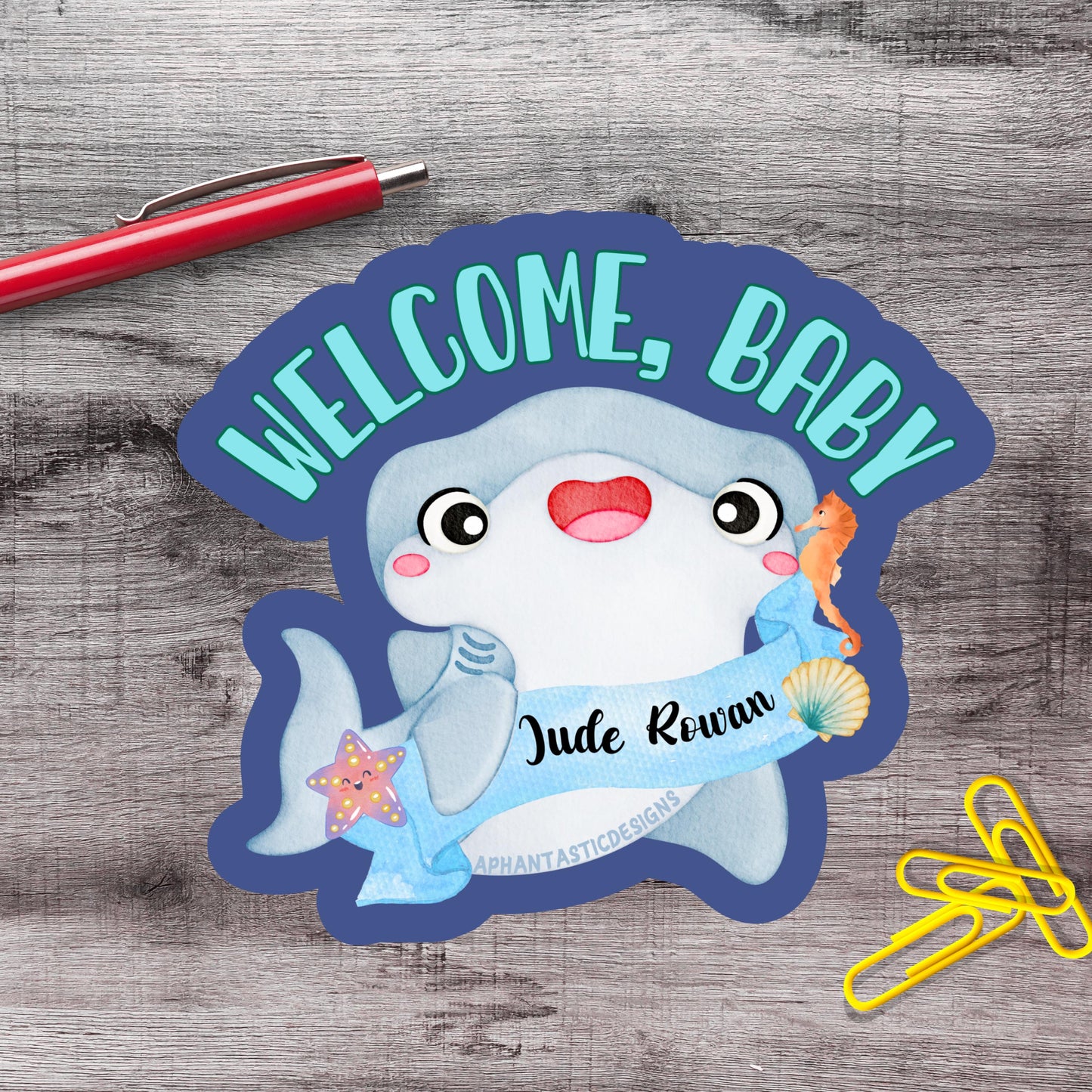 a welcome sign with a cartoon shark and a name tag, placed on a wooden surface alongside a red pen and yellow paper clips.