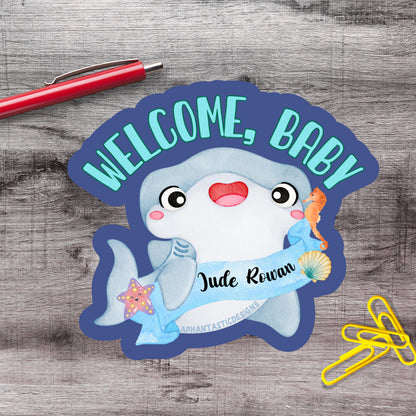 a welcome sign with a cartoon shark and a name tag, placed on a wooden surface alongside a red pen and yellow paper clips.