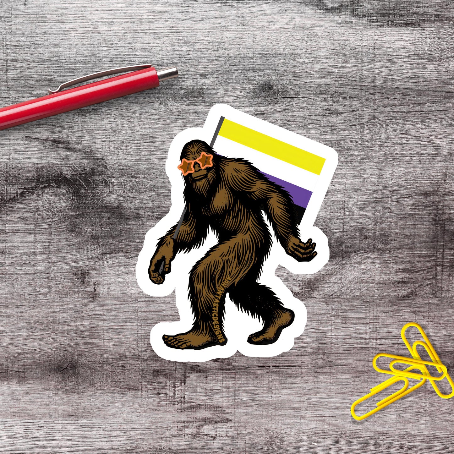 a sticker of a brown, furry creature with a yellow stripe on its back, standing on a wooden surface next to a red pen and a pile of yellow paper clips.