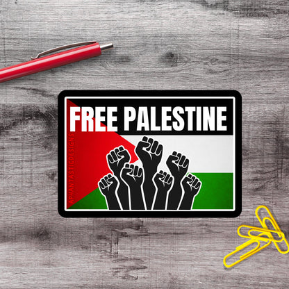a sticker with the text "FREE PALESTINE" and a graphic of raised fists, placed on a wooden surface alongside a red pen and yellow paper clips.