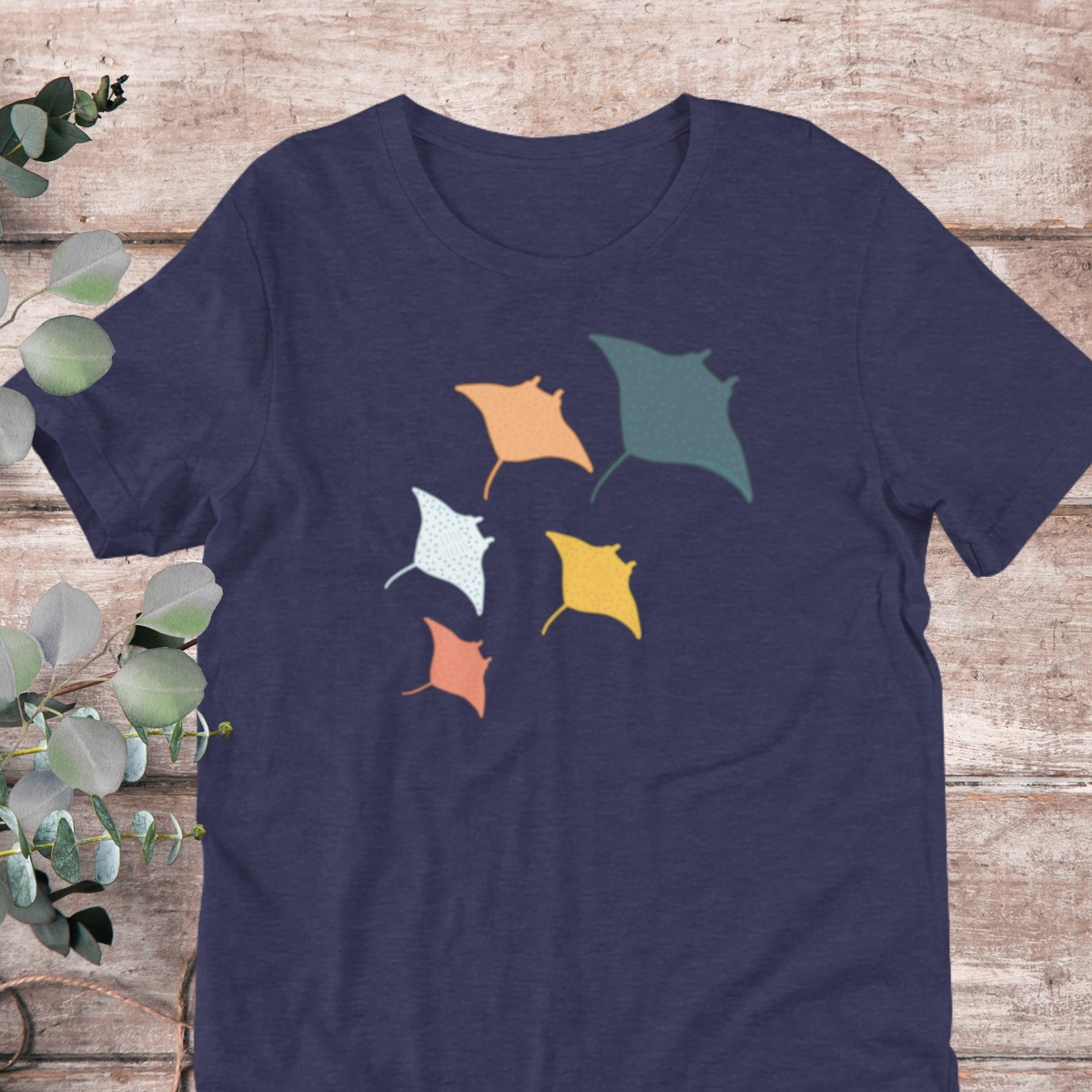A navy blue t-shirt with a graphic design of five colorful stingrays or rays on it, placed on a wooden surface with some greenery in the background.
