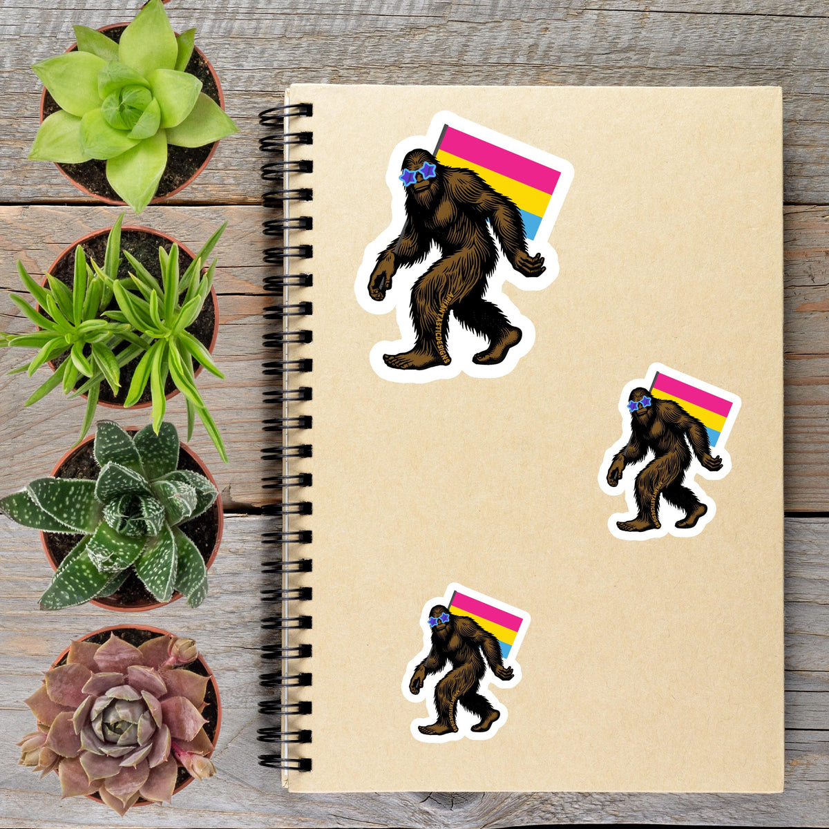 a spiral notebook with stickers of a cartoon Sasquatch wearing a rainbow flag, surrounded by various potted plants on a wooden surface.