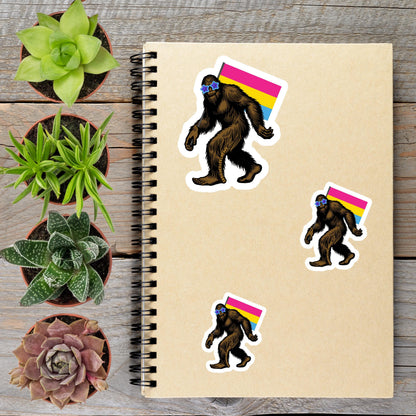 a spiral notebook with stickers of a cartoon Sasquatch wearing a rainbow flag, surrounded by various potted plants on a wooden surface.