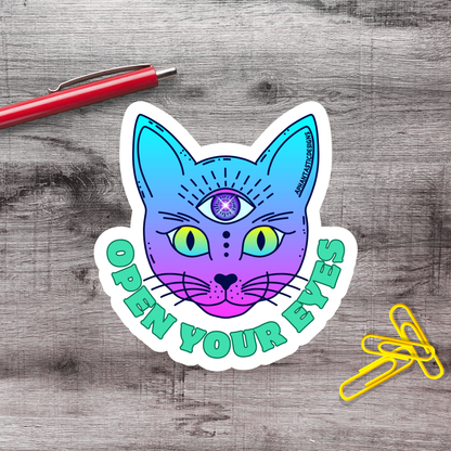 A sticker featuring a colorful cat with an eye design, accompanied by the text "OPEN YOUR EYES" on a wooden surface.