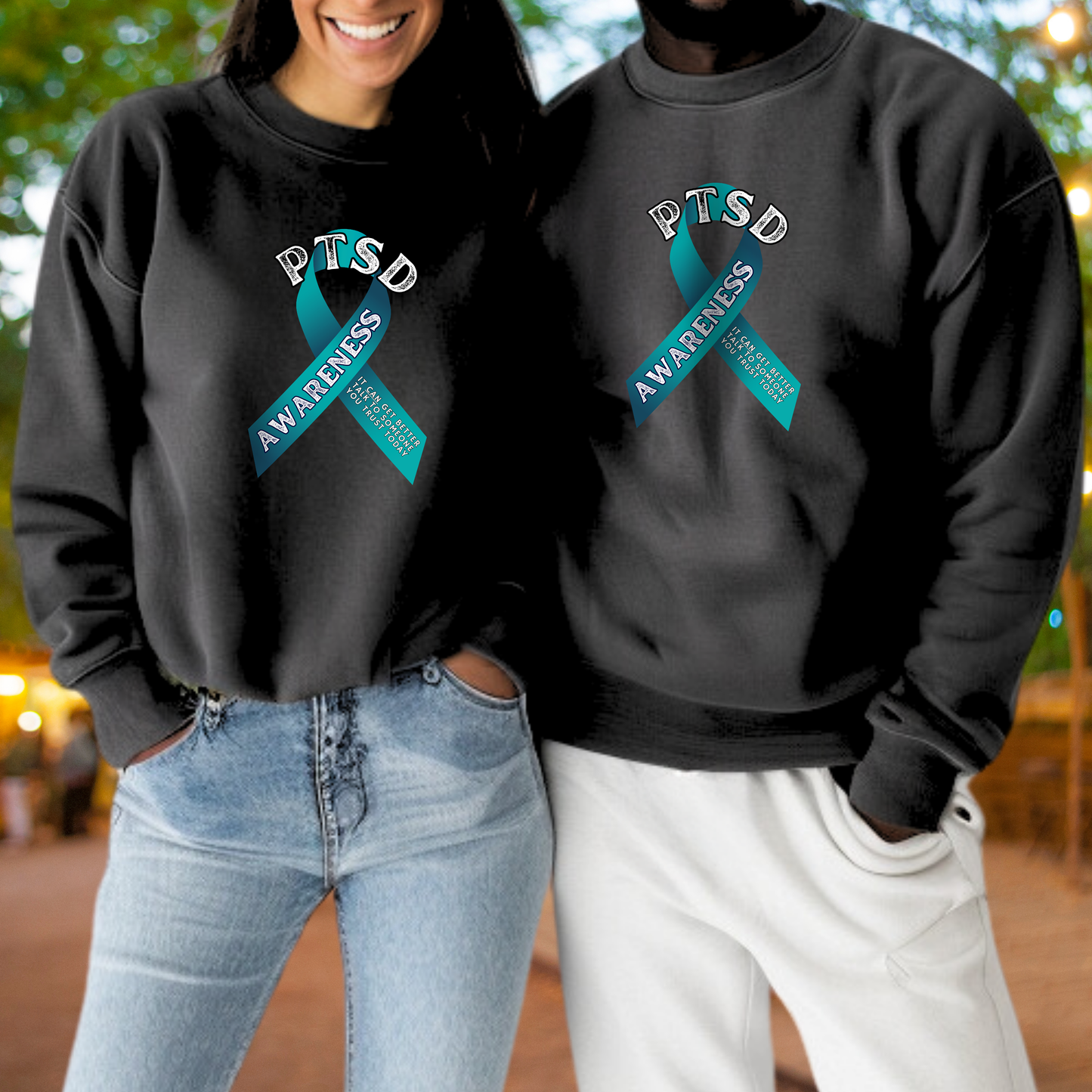 A woman and a man standing side by side, both wearing black sweatshirts with a teal ribbon design and the text "PTSD AWARENESS" printed on them.