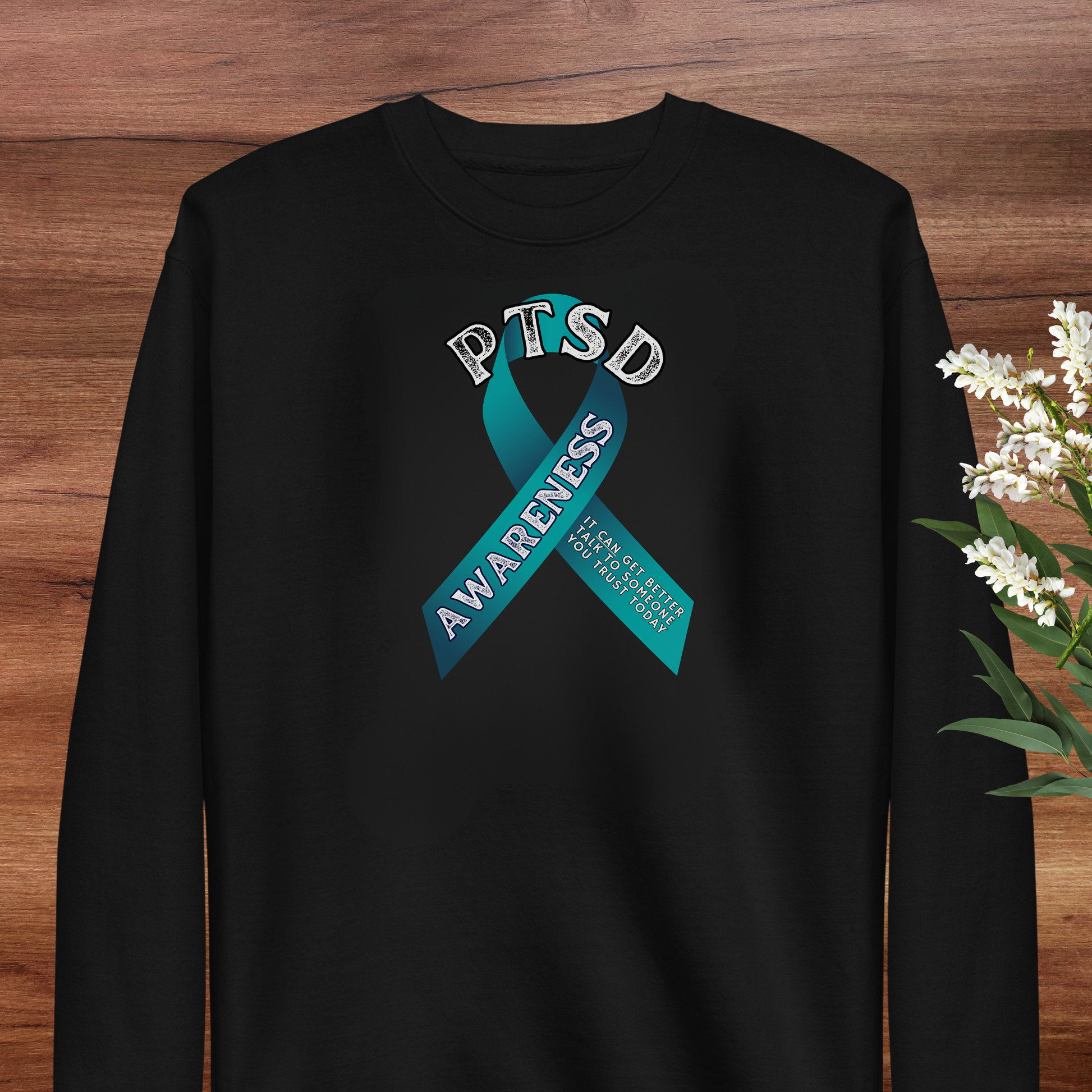 A black long-sleeved shirt with a teal ribbon design that says "PTSD AWARENESS" and "It's not a sign of weakness" printed on it.