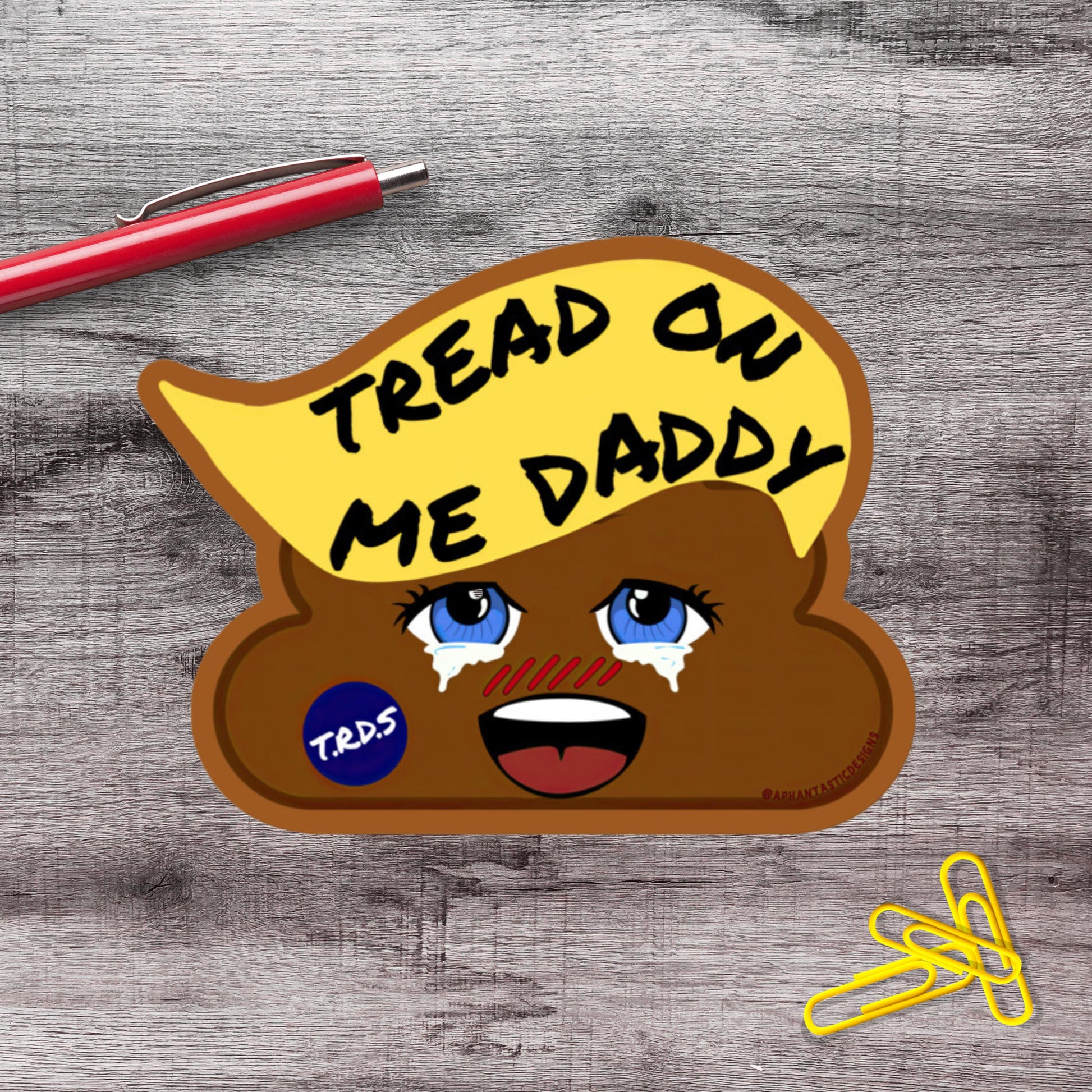 a sticker with a cartoon-like face and the text "TREAD ON ME DADDY" on it, placed on a wooden surface alongside a red pen and some yellow clips.
