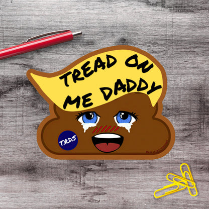 a sticker with a cartoon-like face and the text "TREAD ON ME DADDY" on it, placed on a wooden surface alongside a red pen and some yellow clips.