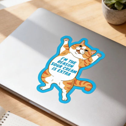 a laptop with a sticker of a cat, a notebook, pens, and a glass of iced coffee on a wooden desk.