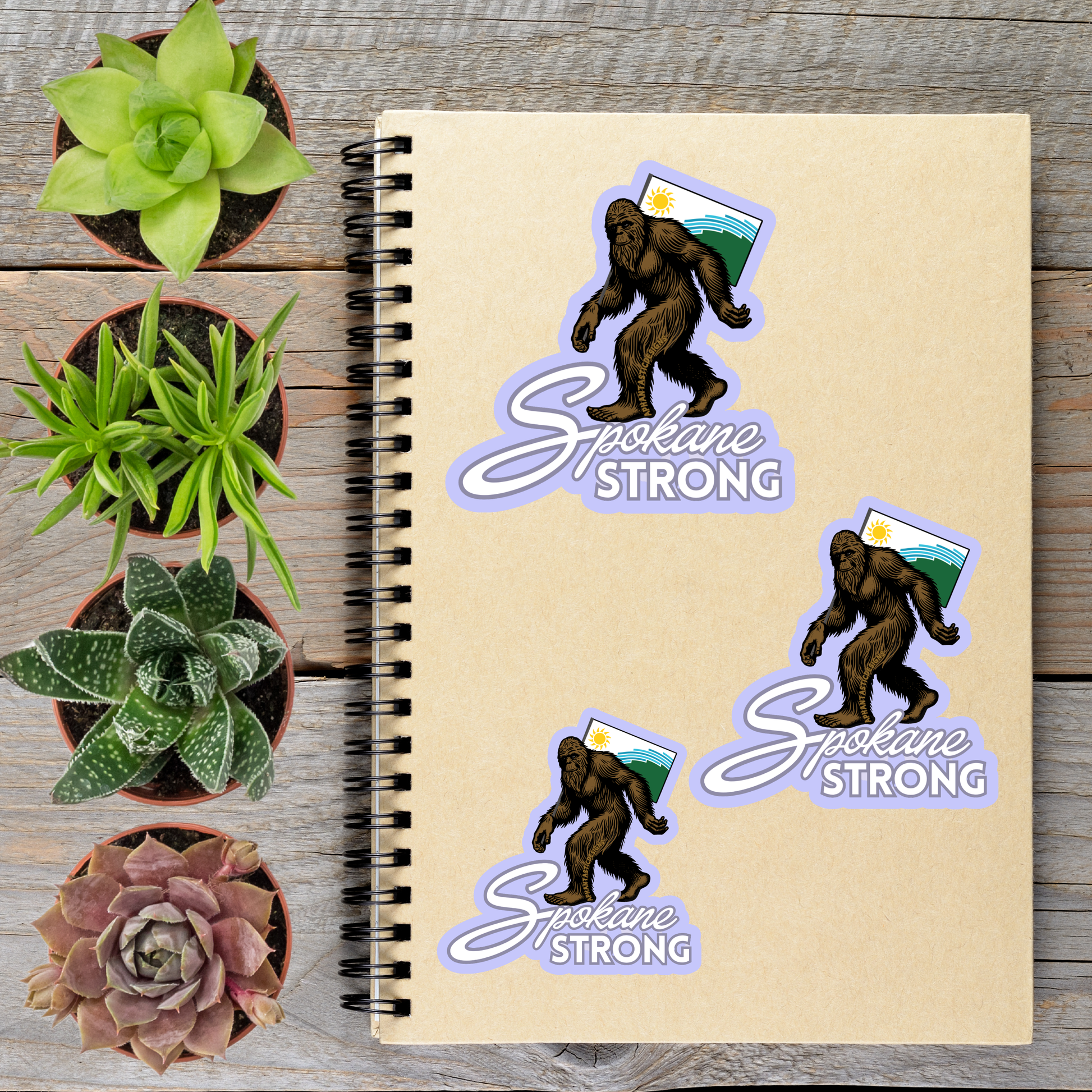 a notebook with stickers of a Sasquatch and the phrase "Sasquatch Strong" on it, along with three potted succulents and a small purple flower.