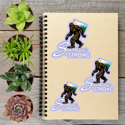 a notebook with stickers of a Sasquatch and the phrase "Sasquatch Strong" on it, along with three potted succulents and a small purple flower.
