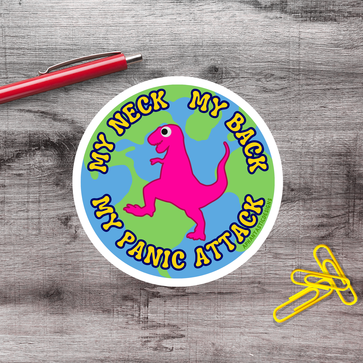 A sticker with a pink dinosaur and the text "My neck, my panic attack" is placed on a wooden surface next to a red pen and yellow paper clips.