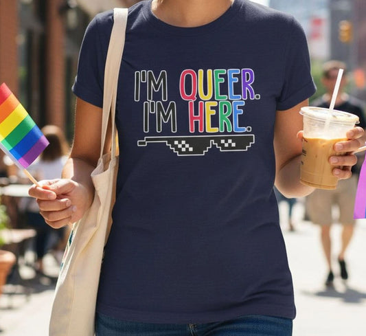 A person wearing a black t-shirt with the text "I'm Queer. I'm Here" printed on it, holding a rainbow flag and a drink.